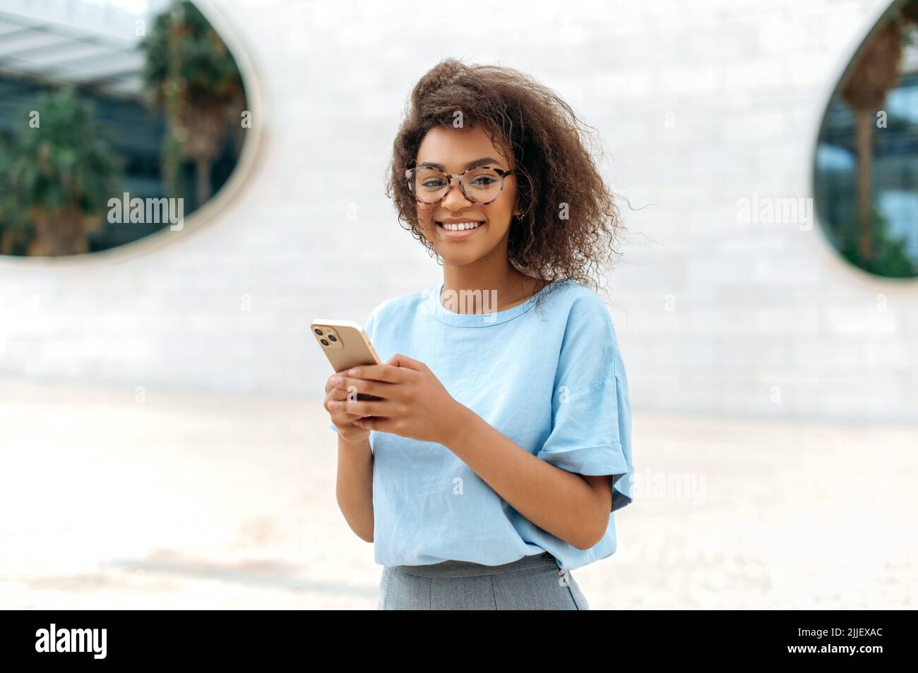 Gadget, drahtlose Technologie. Erfolgreiche, schöne afroamerikanische junge lockige Frau mit Brille, in blauem Hemd, steht im Freien in der Nähe des Geschäftszentrums, nutzt ihr Smartphone, blickt auf die Kamera und lächelt Stockfoto