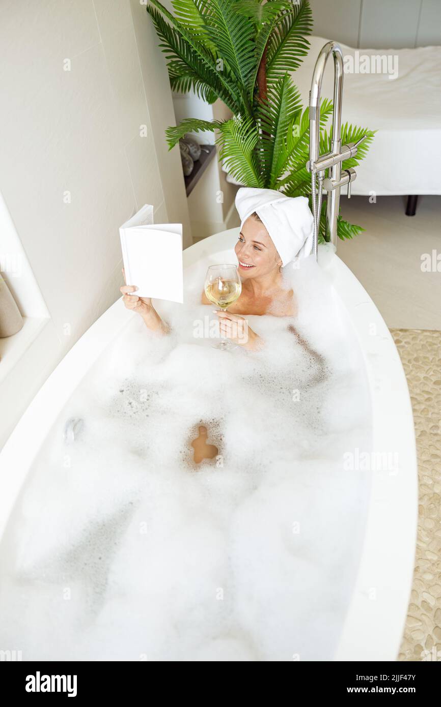 Attraktive kaukasische junge Frau mit Handtuch auf dem Kopf, Baden und Buch mit Wein lesen. Stockfoto