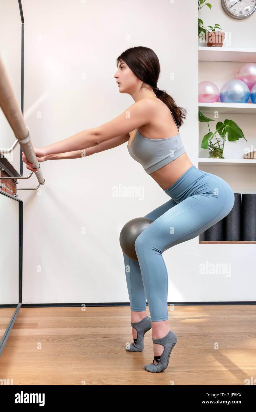 Ganzkörper-Seitenansicht der FIT Ballerina in aktiver Trainingsbekleidung mit Hocke auf Barre und Ball während der Probe im Lichtstudio Stockfoto