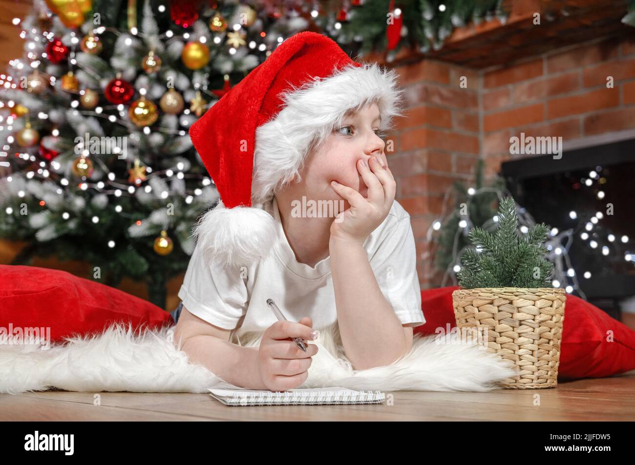 Frohe Weihnachten Kinder. Knabenkind im Weihnachtsmannhut schreibt einen Brief an den Weihnachtsmann unter dem Weihnachtsbaum mit Weihnachtslichtern, in der Nähe des Kamins. Familie Stockfoto