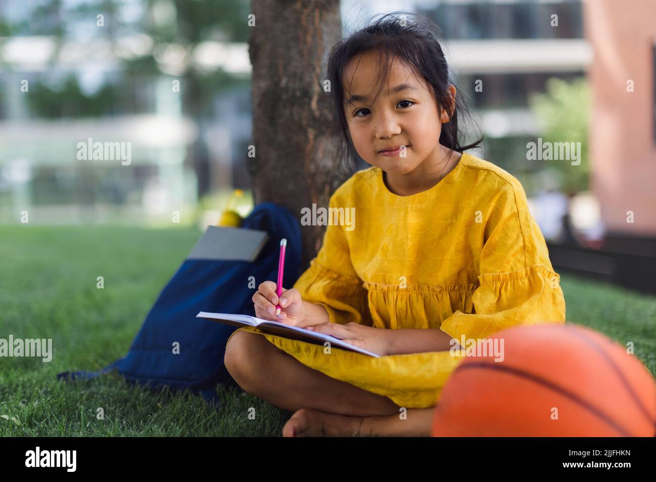 Kleines asiatisches Mädchen, das im öffentlichen Park sitzt und Notizen schreibt. Sommerzeit. Stockfoto