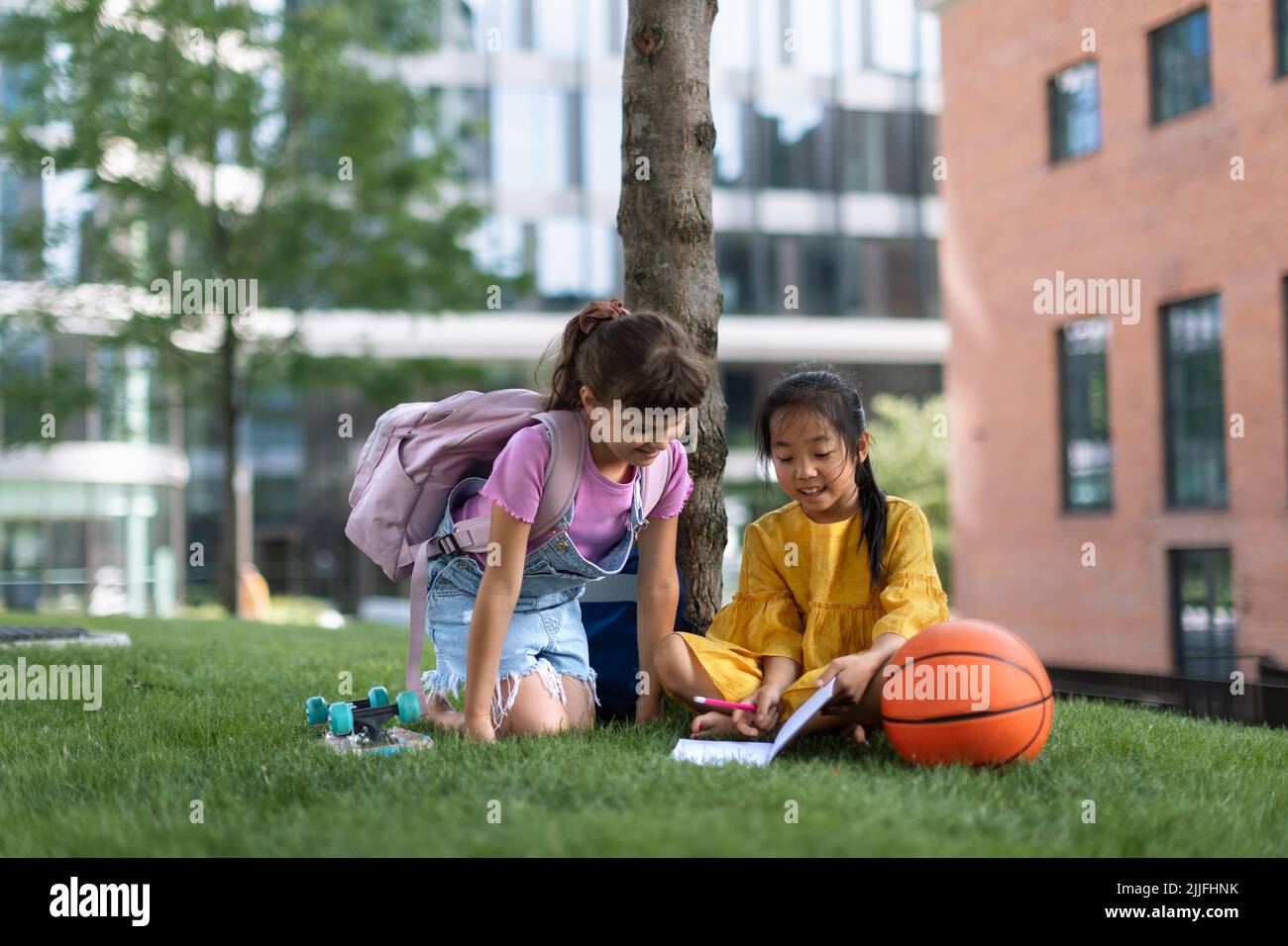 Kleines asiatisches Mädchen mit ihrer Freundin, die im öffentlichen Park sitzt und Notizen schreibt. Sommerzeit. Stockfoto