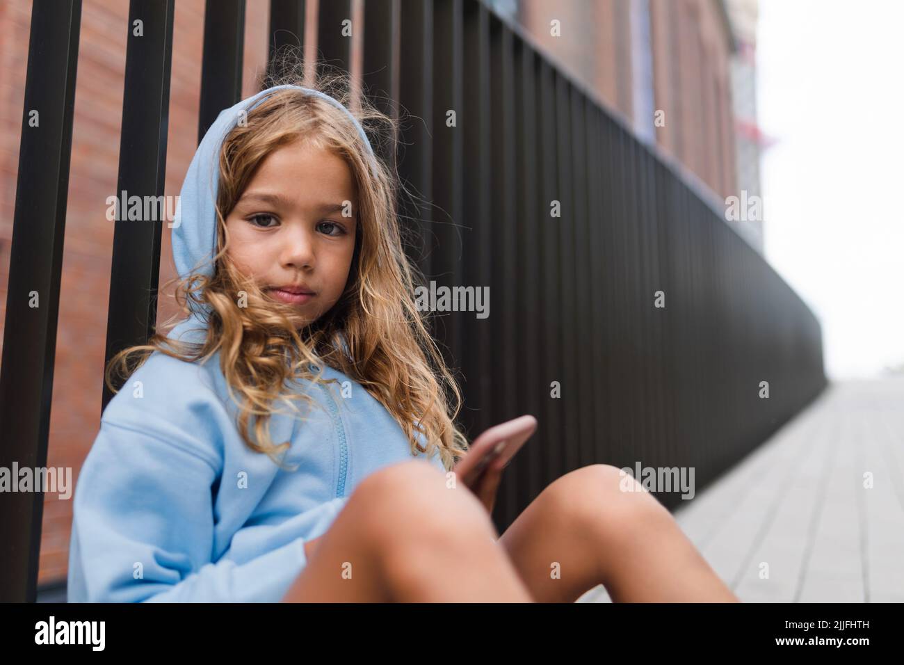 Kleines blondes Mädchen mit Kapuze sitzt auf dem Bürgersteig, hält Smartphone und blickt auf die Kamera. Stockfoto