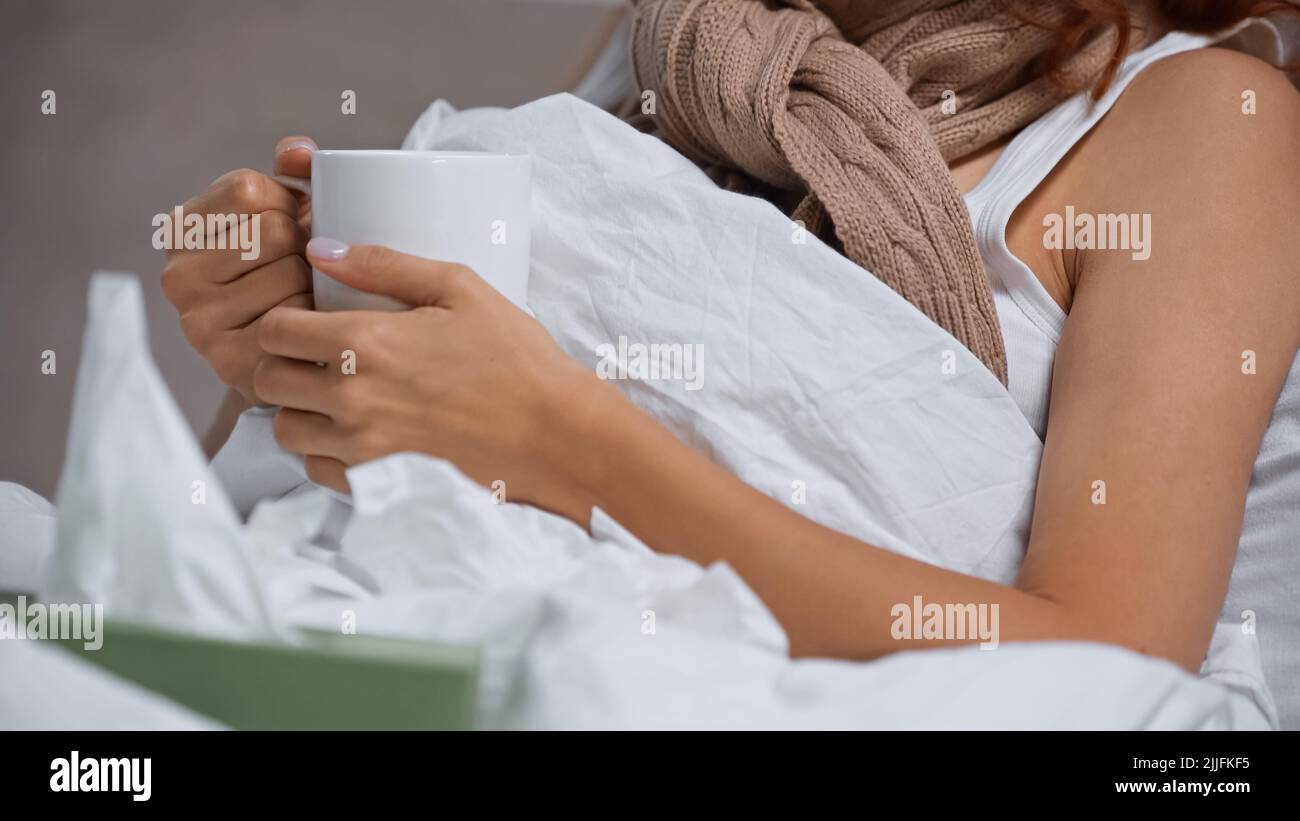 Beschnittene Ansicht einer kranken Frau in einem Schal, der eine Tasse mit einem Getränk in der Nähe von verschwommenen Servietten hält Stockfoto