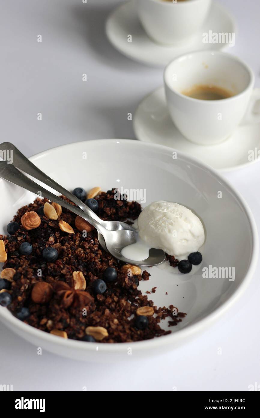 Schokoladengranola Dessert mit Heidelbeeren, Nüssen und Schaufel Vanilleeis auf weißem Tischhintergrund mit Kaffee. Stockfoto