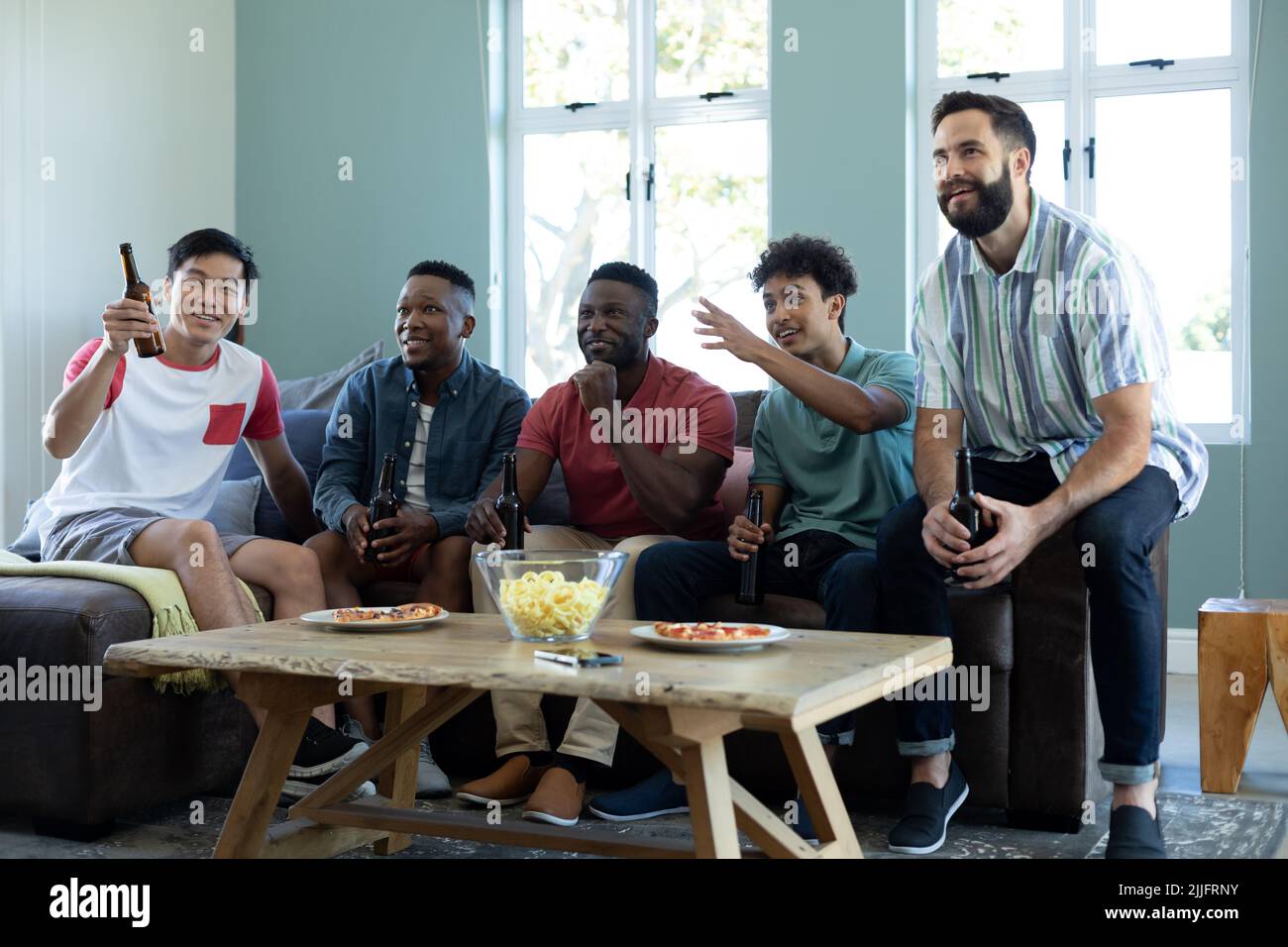 Aufgeregte, multirassische männliche Freunde mit Bierflaschen und Snacks, die sich das Spiel ansehen, während sie sich auf Limonade entspannen Stockfoto