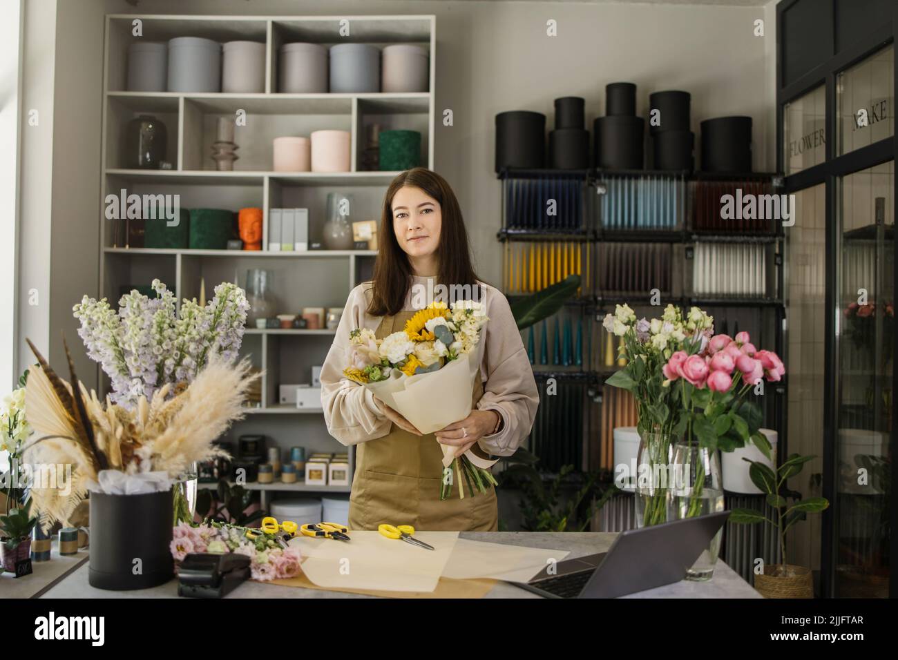 Lächelnder Florist in Uniform in einem Blumenladen hinter dem Tresen mit verschiedenen Blumenarten und riesigem Blumenstrauß in den Händen. Konzept für kleine Unternehmen. Moderne Loft-Innenausstattung. Stockfoto