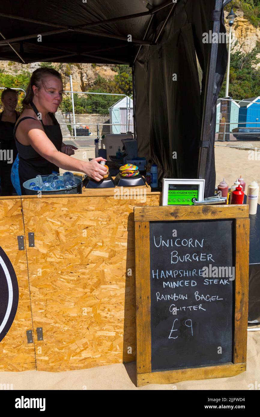 Unicorn Burger Hampshire Hacksteak Regenbogen Bagel Glitzer zum Verkauf £9 auf Food Stand bei der Veranstaltung in Poole, Dorset UK im Juli - Frau sprüht Glitzer auf Stockfoto