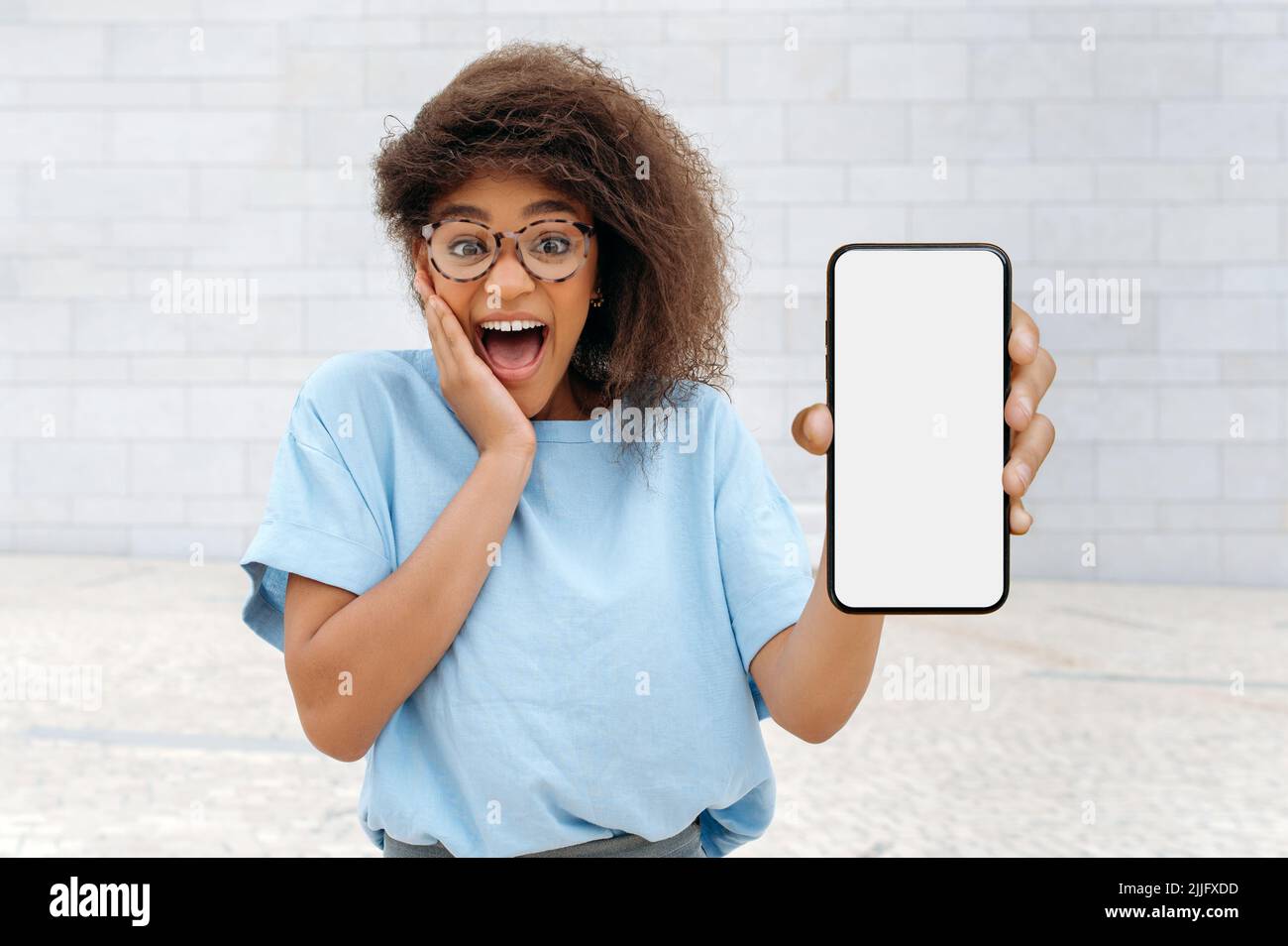 Aufgeregt überrascht freudige afroamerikanische lockige junge Frau, die draußen stand und ihr Smartphone mit weißem Mockup-Bildschirm, Platz für Werbung, Präsentation zeigte, schockiert auf die Kamera aussagt Stockfoto