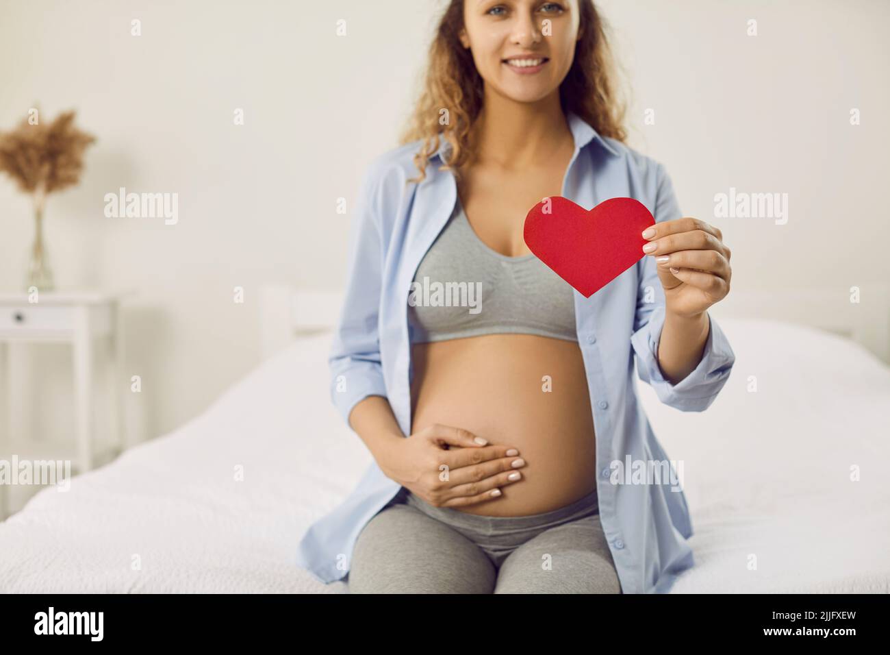Eine glückliche, liebende Schwangerin sitzt auf dem Bett und hält ein Papierherz in den Händen Stockfoto