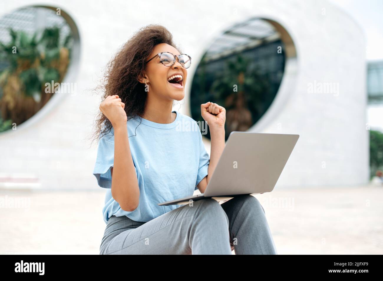 Fröhlich aufgeregt afroamerikanische junge Frau mit Brille, nutzt Laptop, während sie im Freien sitzt, die Hände vor Aufregung hebt, Fäuste streicht, Erfolg feiert, gewinnt, glücklich lächelt. Gute Nachrichten, gewinnen Stockfoto