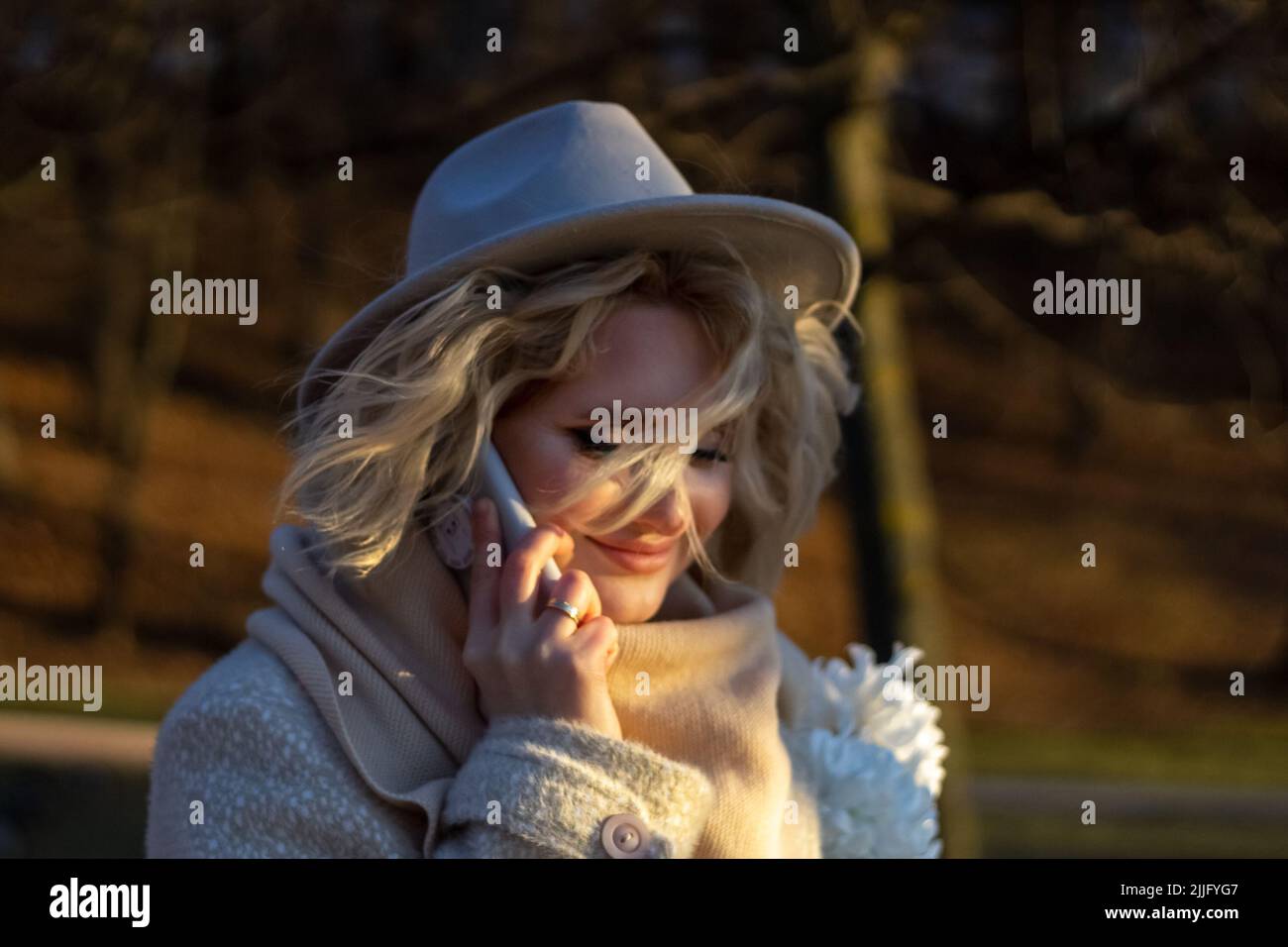Eine Frau in einem Hut mit einem flatternden Haarsträhnen telefoniert in einem Herbstpark bei Sonnenuntergang. Sie ist glücklich und lächelt. Weicher Fokus. Spot. Ein geldautomat Stockfoto
