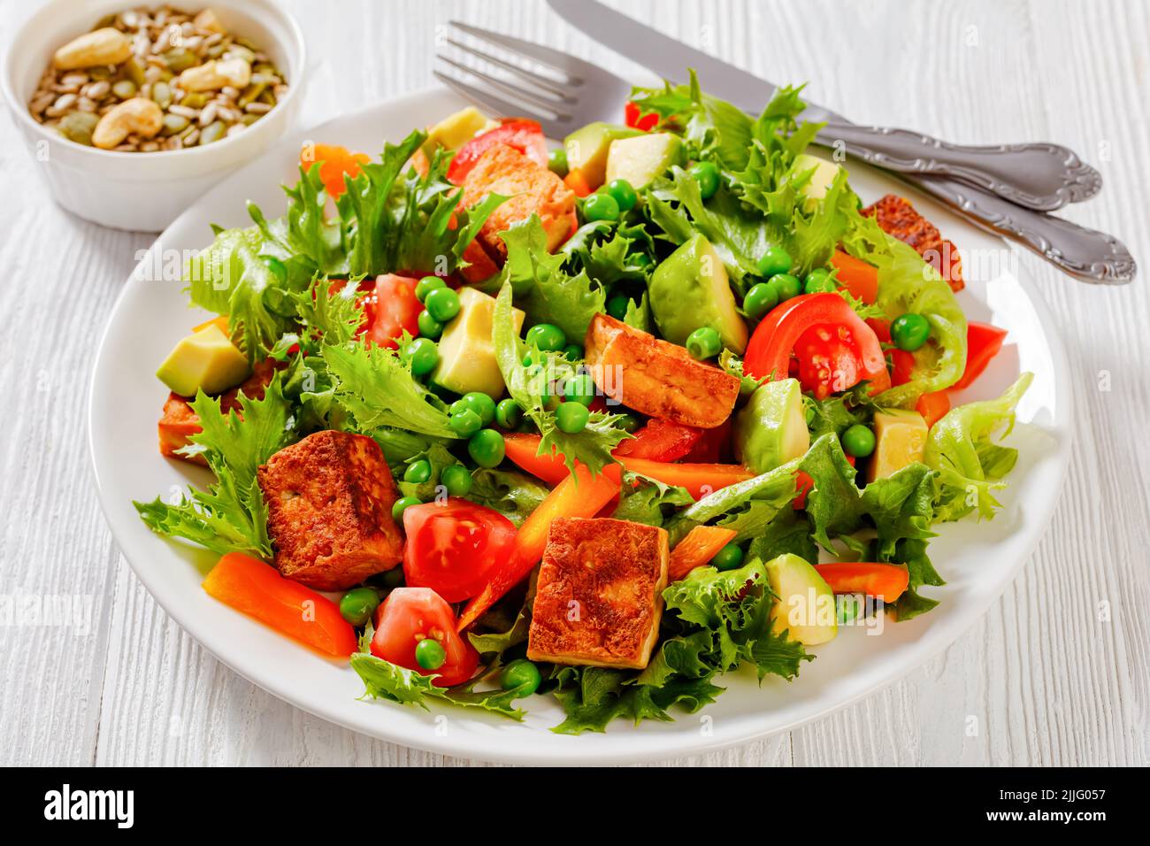 Tofu-Salat mit Avocado, knackigen Salatblättern, grünen Erbsen, Tomaten, gelber Paprika auf weißem Teller, horizontale Ansicht von oben Stockfoto