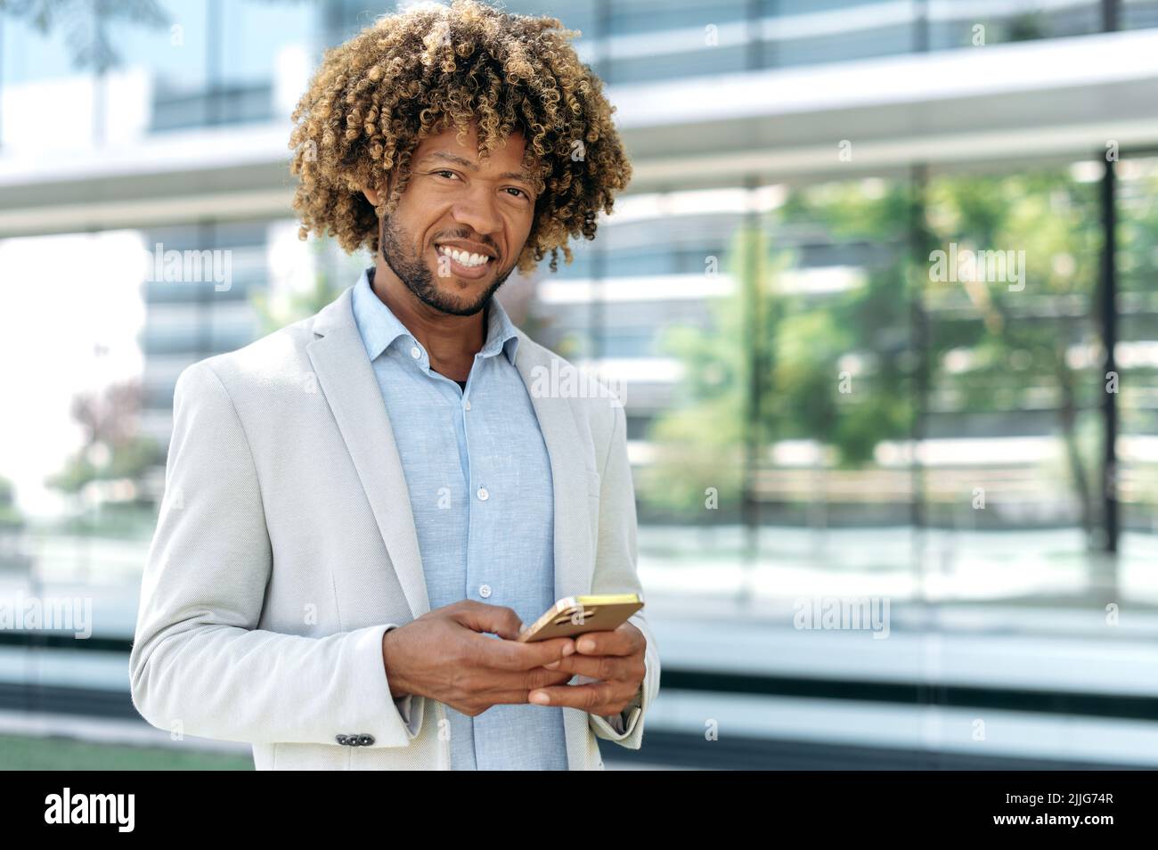 Erfolgreicher, selbstbewusster, eleganter brasilianischer lockiger Mann, der stilvolle formelle Kleidung trägt, steht im Freien in der Nähe des Geschäftszentrums, nutzt sein Smartphone, blickt auf die Kamera und lächelt. Gadget, drahtlose Technologie Stockfoto