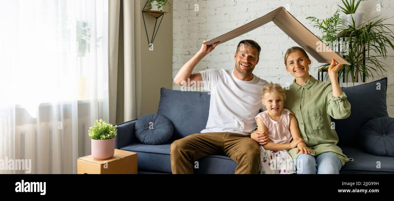 Junge Familie mit Kind sitzt auf der Couch und hält Pappdach über Kopf. Umzug in neues Zuhause, Hypothek und Versicherungskonzept. Platz kopieren Stockfoto