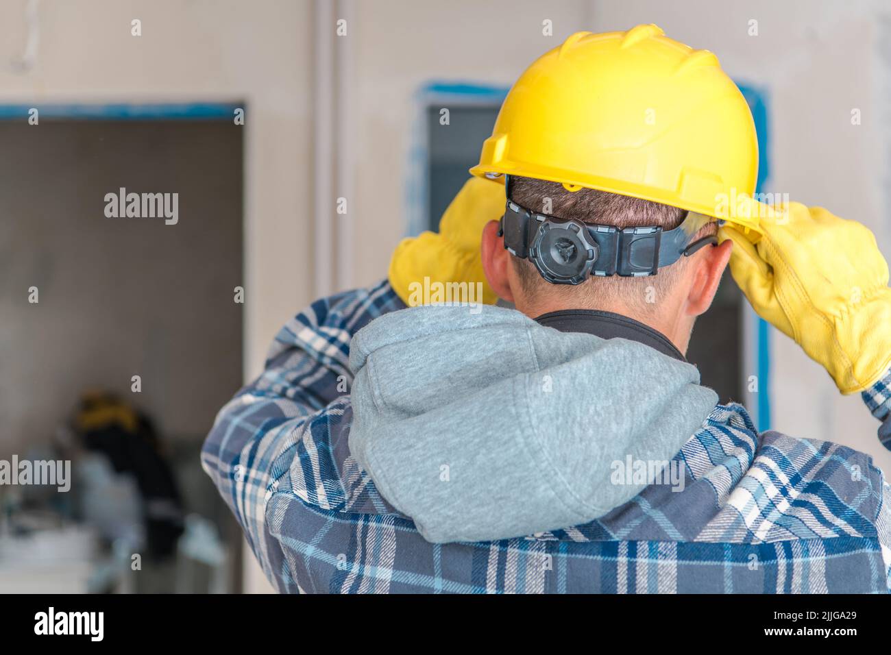 Kaukasischer Bauarbeiter, der seinen verstellbaren gelben Sicherheitshelm aufsetzt, bevor er eine Baustelle begibt, um seine Arbeit zu beginnen. Rückansicht. Stockfoto