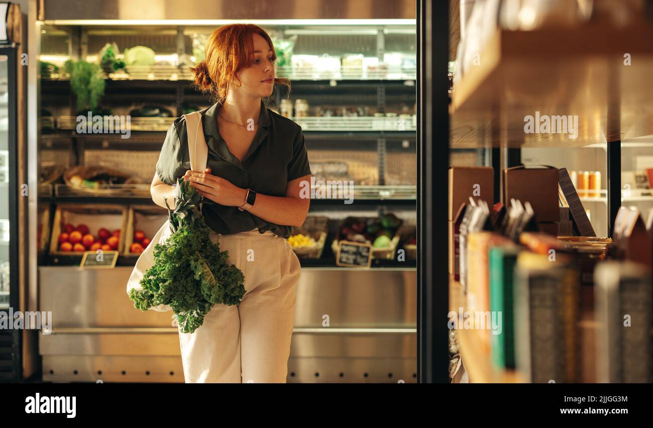 Weibliche Kundin schaut durch ein Lebensmittelregal, während sie eine Tasche mit frischem Bio-Gemüse in einem Lebensmittelgeschäft trägt. Junge Frau macht etwas Lebensmittelgeschäft sho Stockfoto