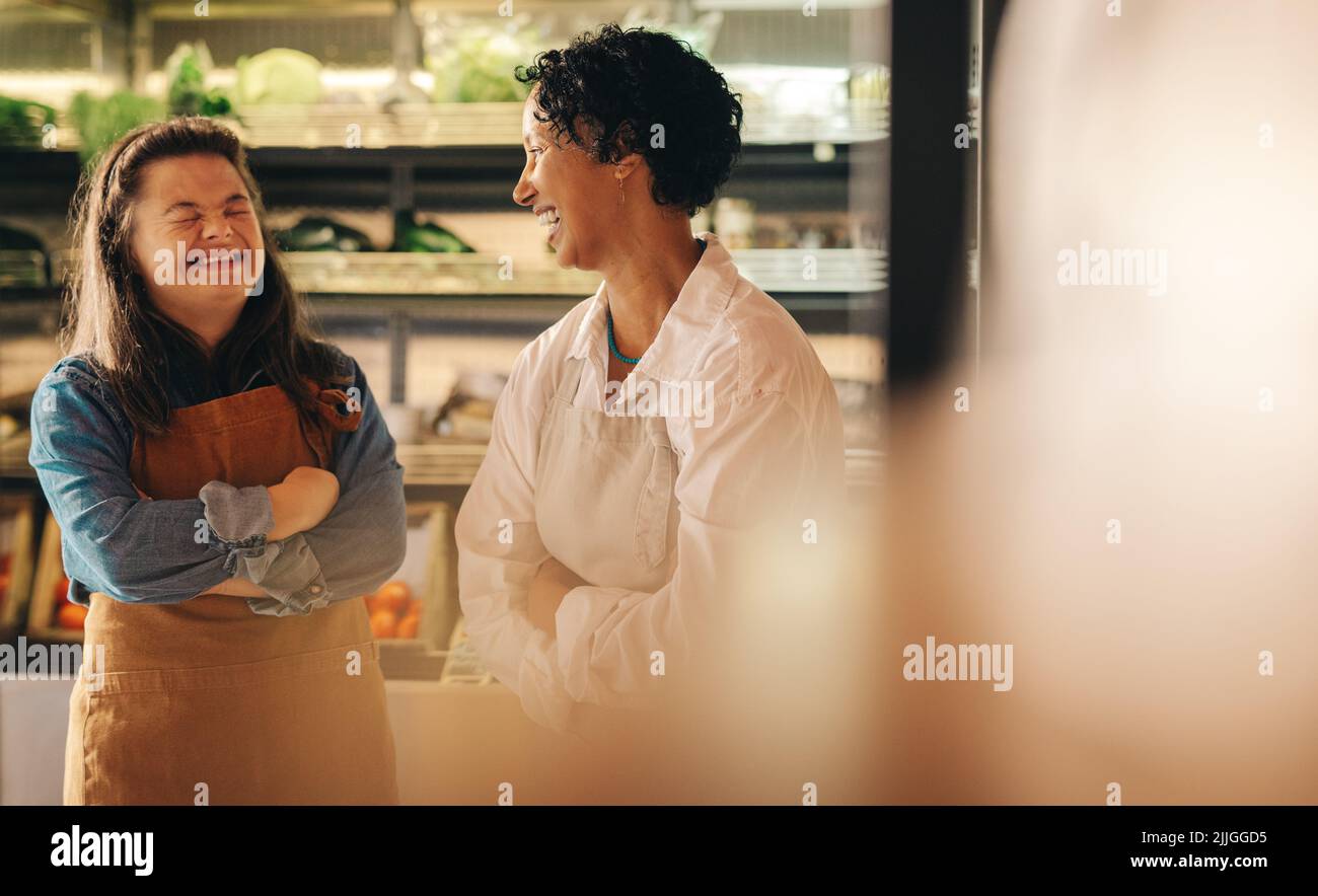 Lebensmittelarbeiter lachen fröhlich, während sie in ihrem Supermarkt zusammen stehen. Glückliche Frau mit Down-Syndrom, die ein kleines Unternehmen mit h Stockfoto