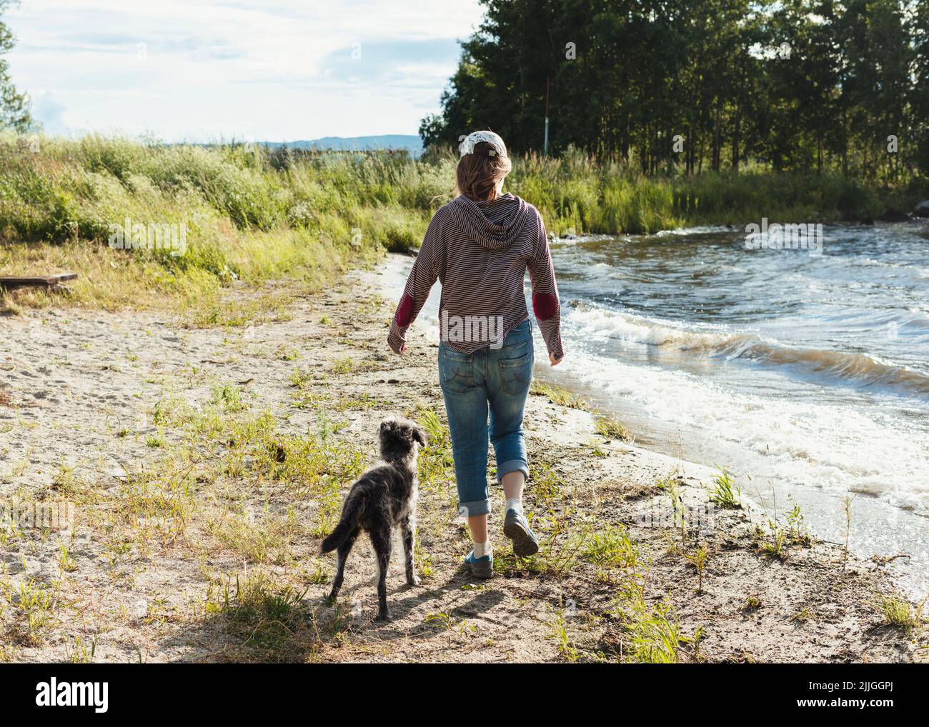 Rückansicht einer jungen Frau, die am Seeufer entlang geht, mit Hundereisen und Wanderungen mit Haustieren Stockfoto