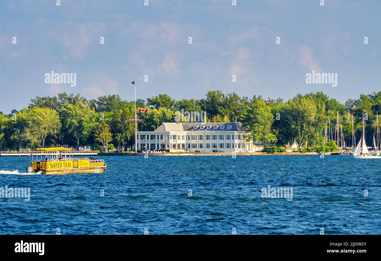 Gebäude des Royal Canadian Yacht Club auf ward's Island of Toronto Islands, mit einem gelben Wassertaxi auf blauem Wasser des Innenhafens. Stockfoto