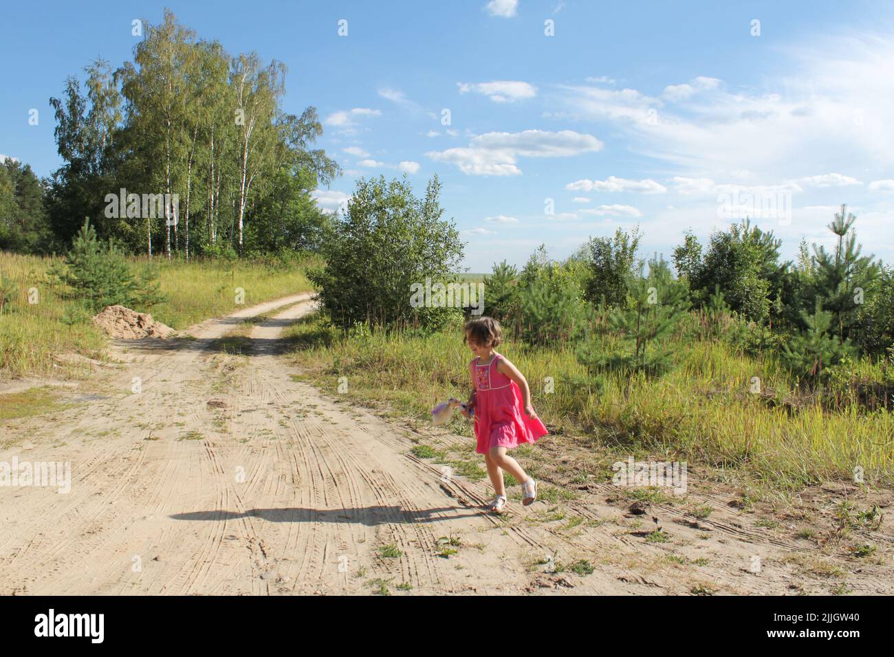 Cutie Kleinkind Mädchen in rosa Kleid mit einer Puppe läuft auf sandigen Weg zwischen Feldern und Wald. Speicherplatz kopieren. Stockfoto