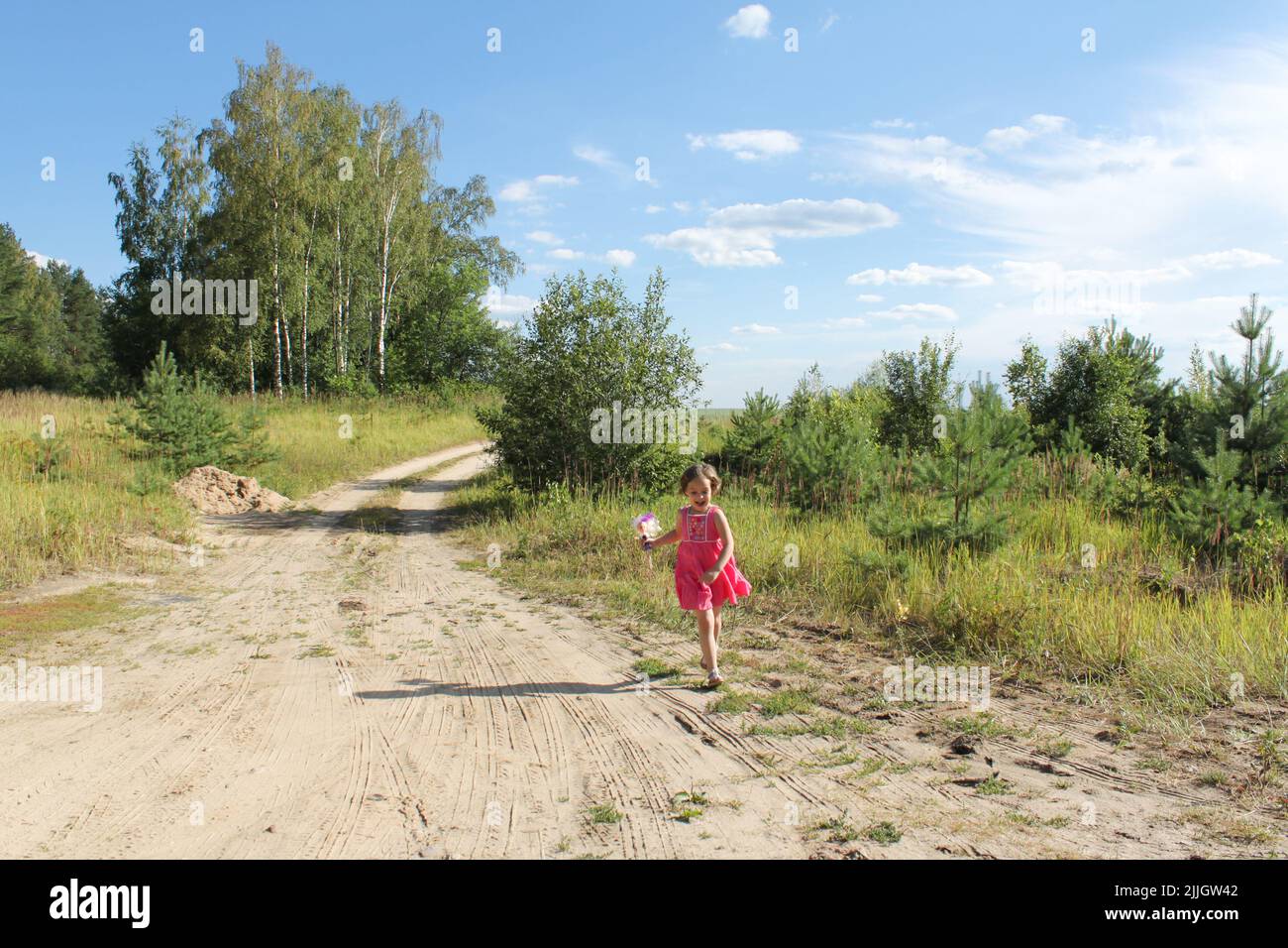 Lustige Kleinkind Mädchen in rosa Kleid mit einer Puppe läuft auf sandigen Weg zwischen Feldern und Wald. Speicherplatz kopieren. Stockfoto
