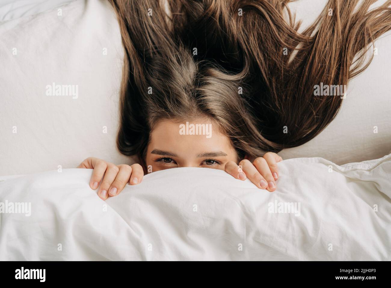 Am Morgen guckt eine junge Frau unter der Decke, die auf dem Bett liegt, heraus. Stockfoto