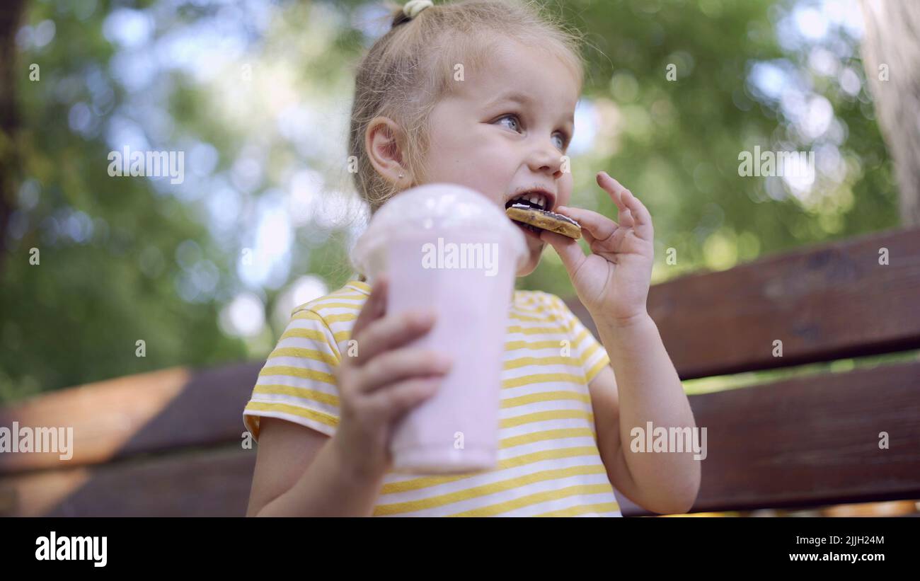 Das kleine Mädchen isst einen bunten Lebkuchen und hält einen Milchshake in der Hand. Nahaufnahme von niedlichen Mädchen auf Parkbank sitzen und essen Cookies mit einem Stockfoto
