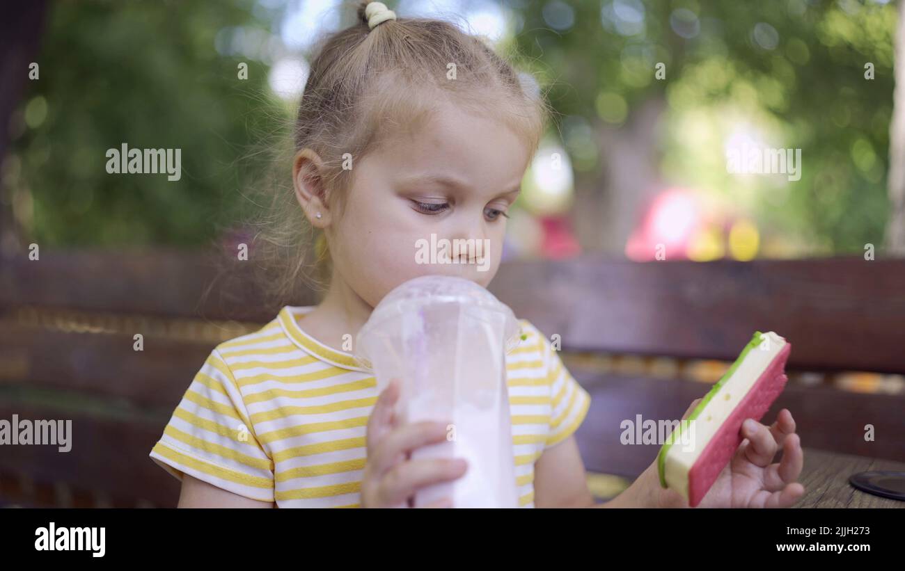 Das kleine Mädchen trinkt einen Milchshake durch einen Strohhalm und schmunkt in der anderen Hand an Eis. Nahaufnahme Porträt von niedlichen Mädchen auf Parkbank A sitzen Stockfoto