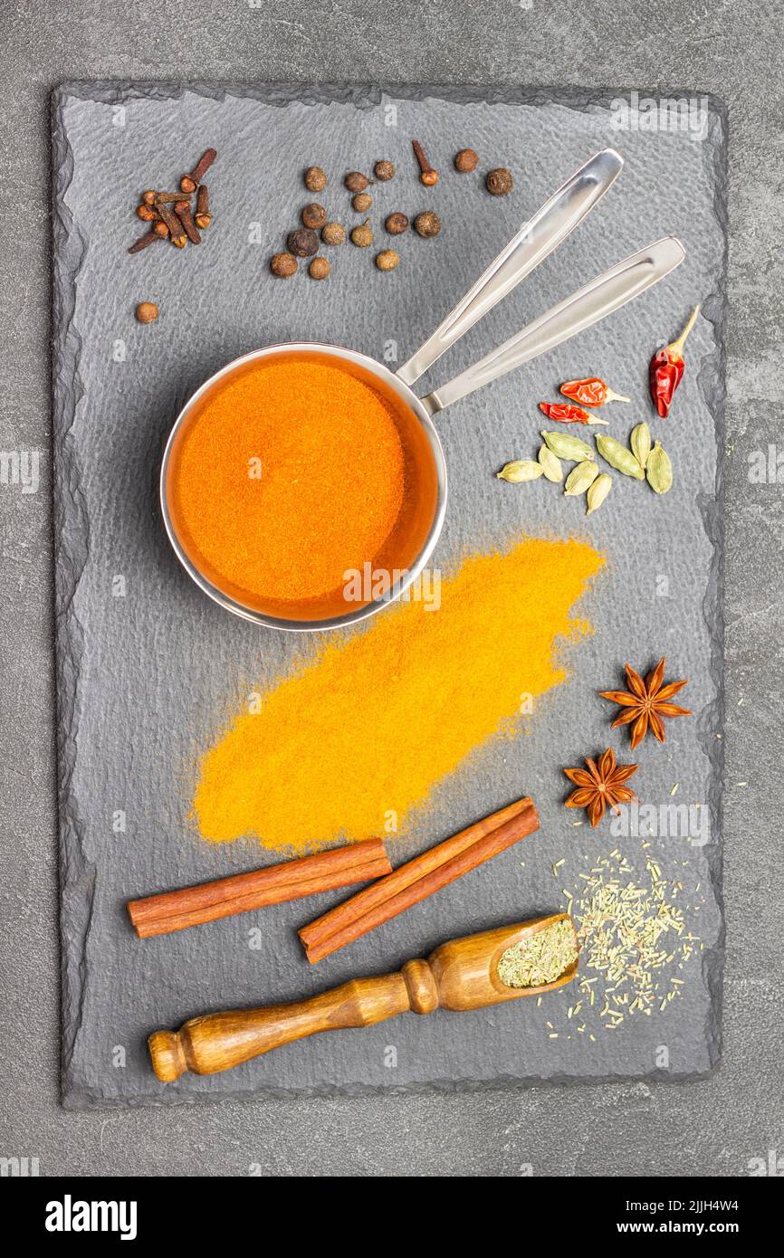 Gemahlener Paprika in Metallschale, gemahlener Kurkuma, Zimtstangen und trockenes Kraut in Schaufel auf Steinbrett. Flach liegend. Schwarzer Hintergrund Stockfoto