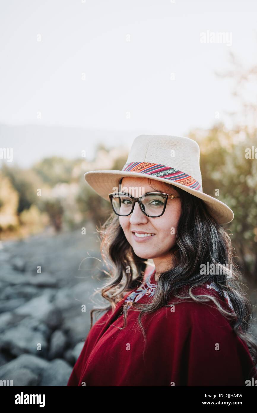 Lächelnde lateinische Frau, die einen traditionellen roten Poncho, eine Brille und einen Hut in einem natürlichen Raum bei Sonnenuntergang trägt. Vertikal Stockfoto