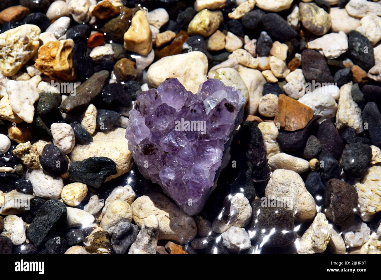 Wunderschöne violette Amethyst-Kristalle auf feuchten Kieselsteinen am Meer. Leuchtend lila Amethyst Druse in Form eines Herzens auf dem Hintergrund des Meeres. Stockfoto