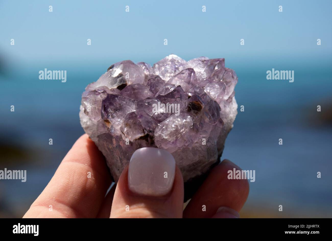 Schöne lila Amethyst Kristalle in einer weiblichen Hand. Druse Amethyst in Form eines Herzens auf dem Hintergrund des Meeres. Stockfoto