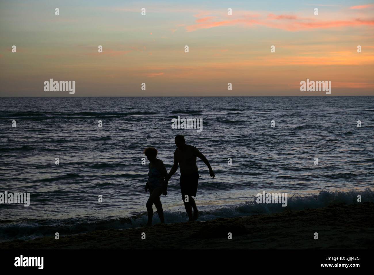 Silhouette eines alten Paares, das bei Sonnenuntergang gegen dramatische rote Meereswellen mit sanftem, dunklem Wasser am Abend läuft Stockfoto