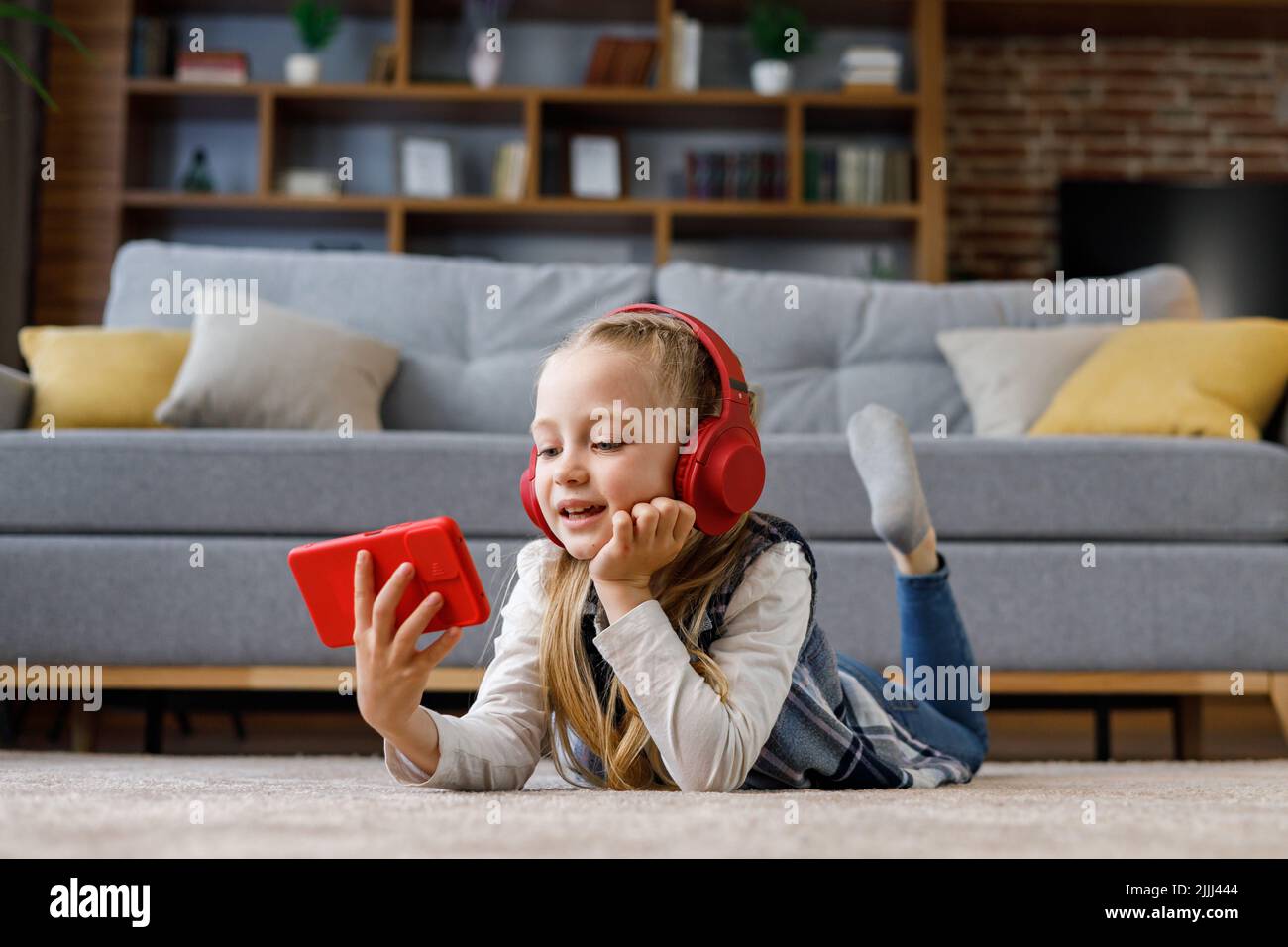 Kleines Mädchen mit roten Ohrhörern, das zu Hause auf dem Teppich liegt. Niedliches lächelndes Kind, das Smartphone hält, Cartoons gucken, im Internet surfen oder zusehen Stockfoto