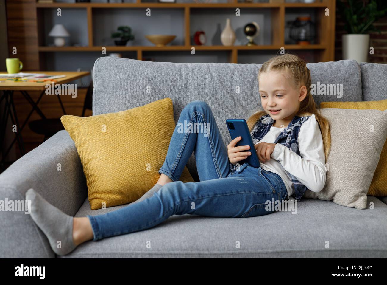 Porträt von niedlichen kleinen niedlichen Mädchen hält Smartphone und ruht auf dem Sofa zu Hause. Auswählen von Lieblingsmusik oder -Cartoons, SMS senden, Surfen Stockfoto