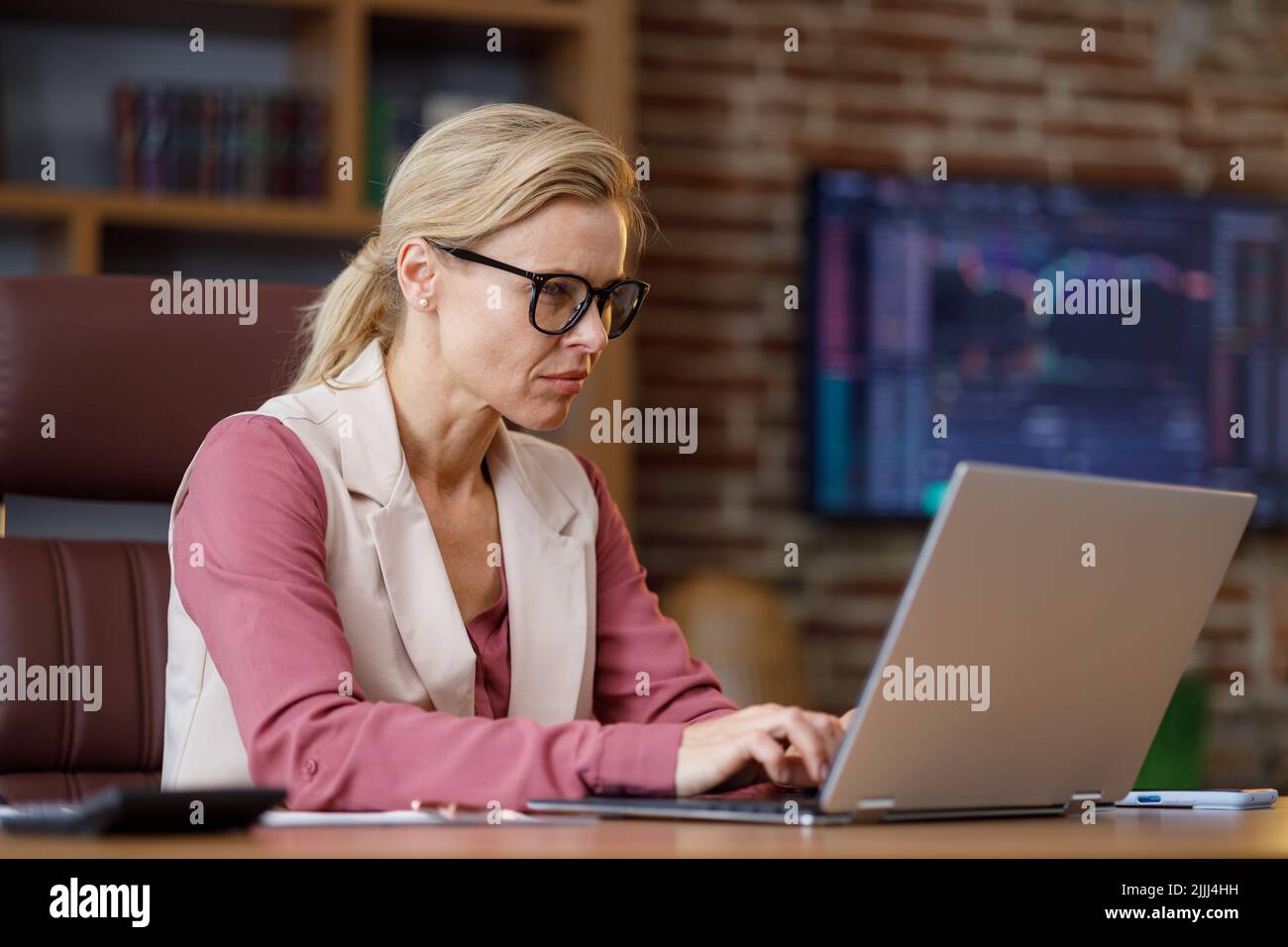 Porträt einer erwachsenen Geschäftsfrau, die im Büro mit einem Laptop arbeitet. Weibliche Trader schreiben Nachrichten und machen Papierkram mit Börsengraphen im Hintergrund Stockfoto