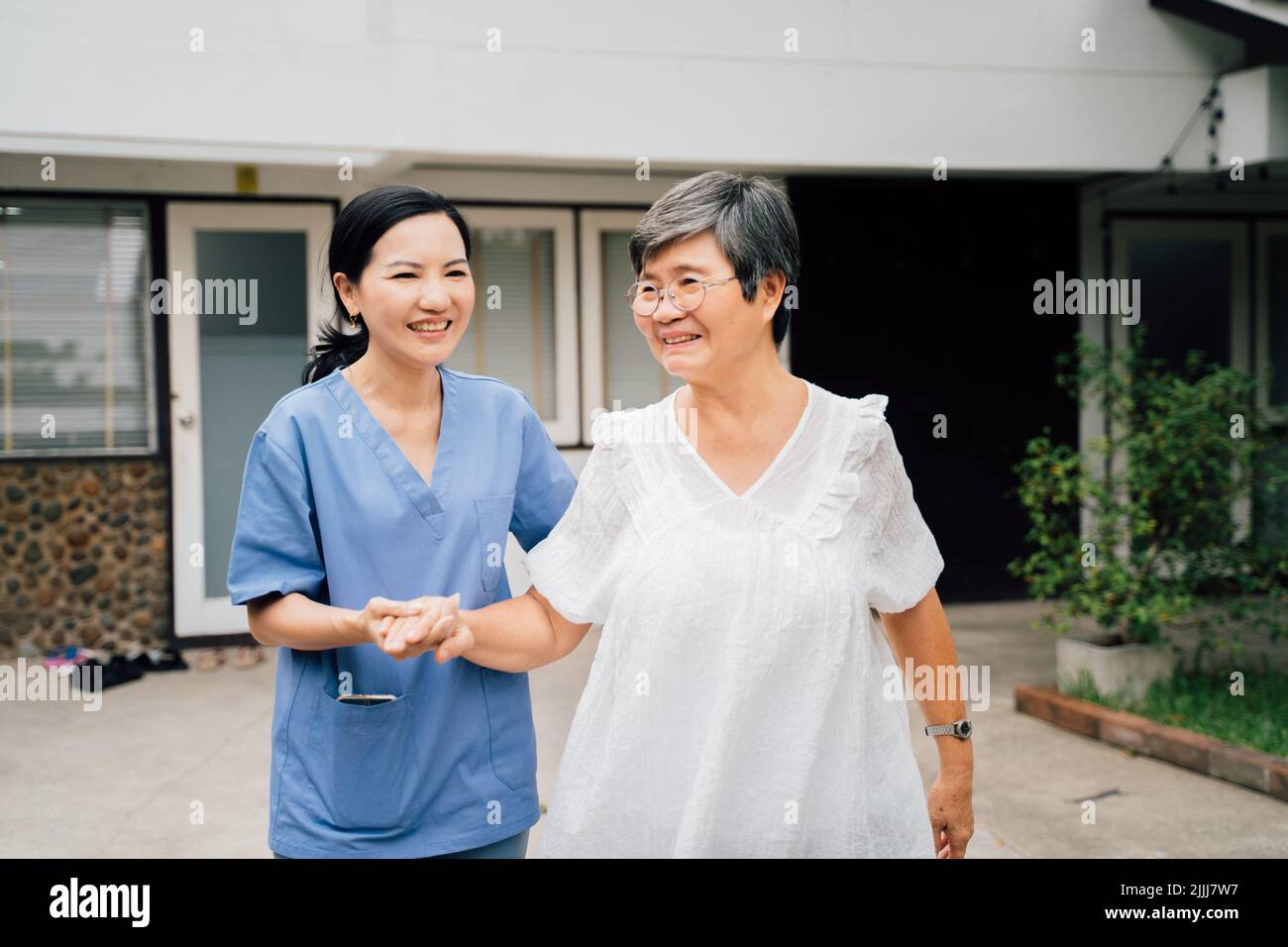 Optimistische Krankenschwester in blauer Uniform, die eine ältere asiatische Frau unterstützt, während sie gemeinsam durch das Tor vor dem Haus geht Stockfoto