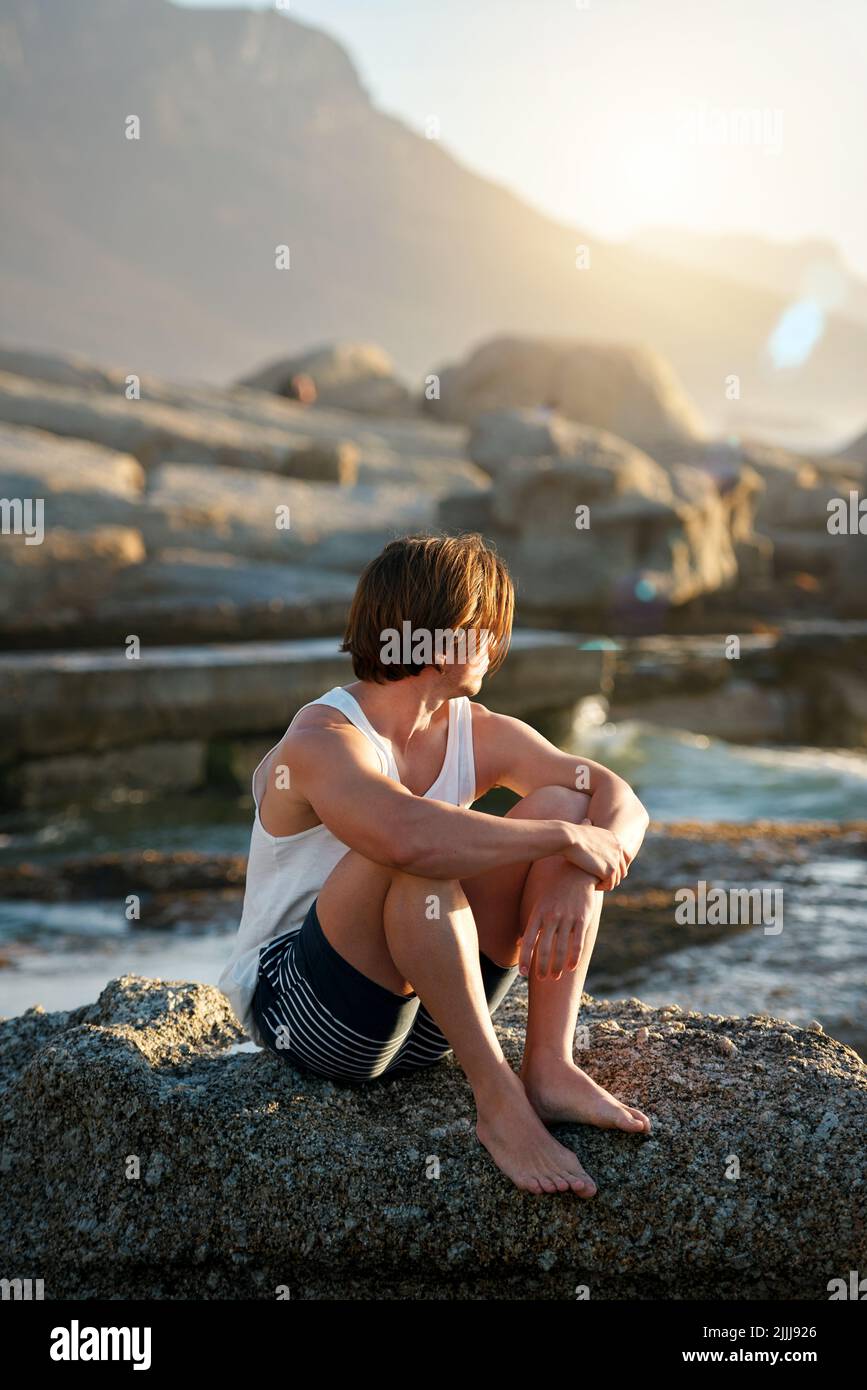 Junger Mann, der am Strand sitzt und den Sonnenuntergang anschaut und dabei den Ozean beobachtet Stockfoto