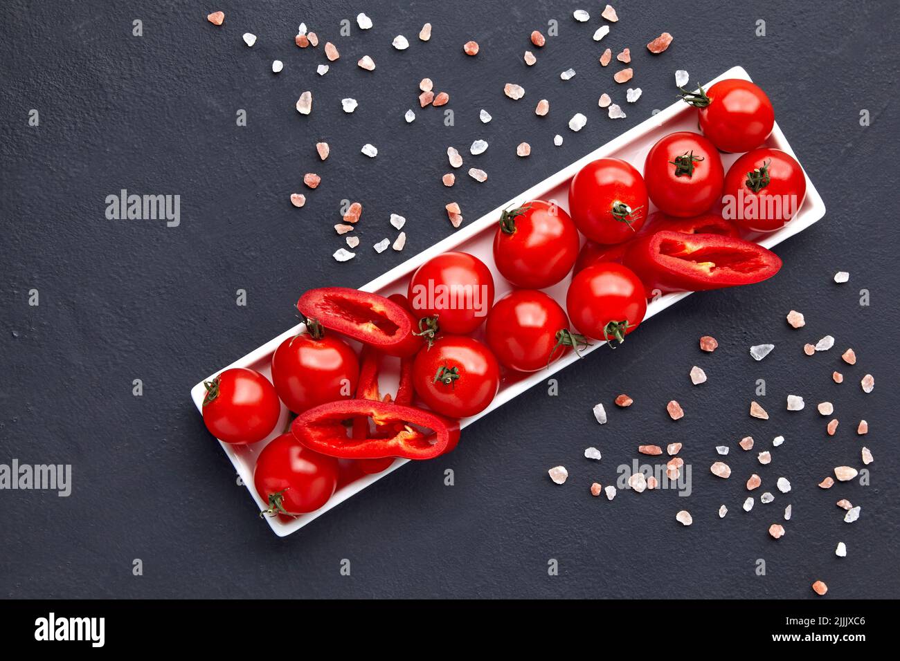 Frisch gewaschene Kirschtomaten und Paprikascheiben auf einer weißen länglichen Schüssel mit rosa Salz auf einem schwarzen Betontisch Stockfoto