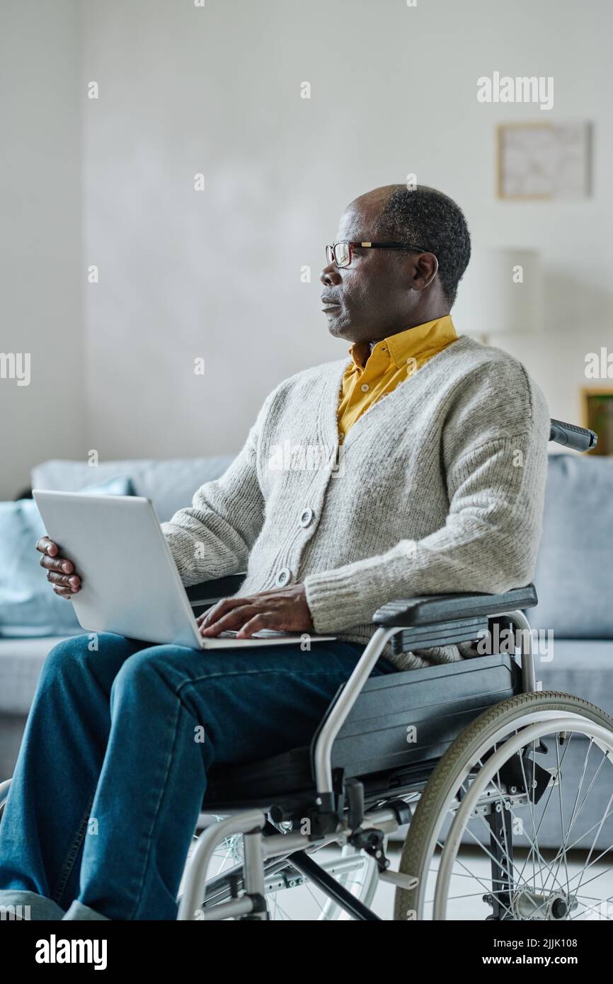 Afrikanischer reifer Mann, der auf dem Rollstuhl sitzt und mit nachdenklichen Worten wegschaut, während er zu Hause mit einem Laptop online arbeitet Stockfoto