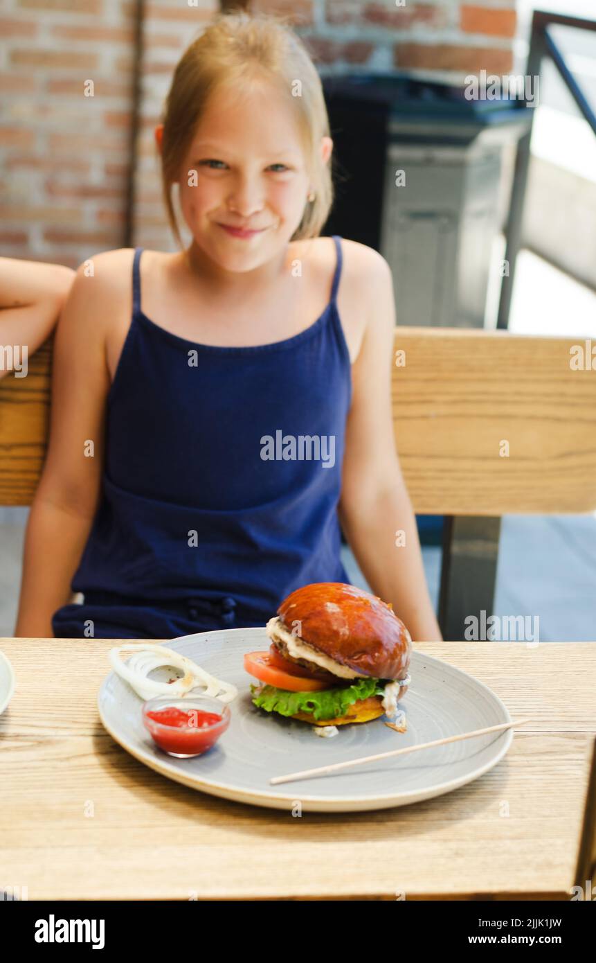 Fröhliches Mädchen, Teenager, die in einem Restaurant auf einer Sommerterrasse einen Burger mit Fleischpastete essen. Essen und Lifestyle. Fast Food. Mittagessen Stockfoto