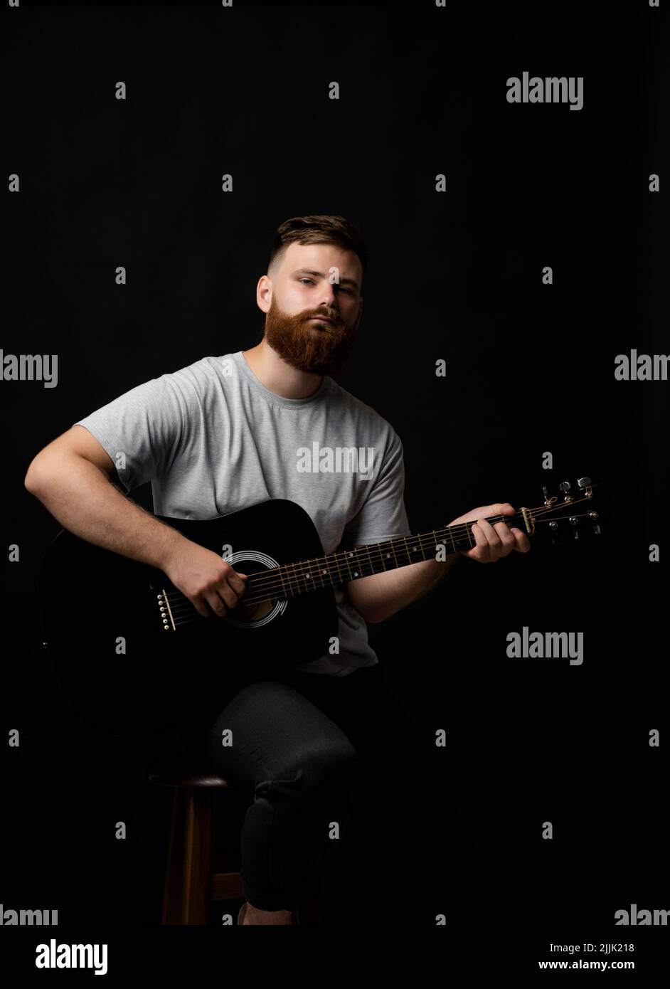 Bärtiger brutaler Gitarrist spielt in einem schwarzen Raum eine akustische Gitarre und schaut in die Kamera. Das Konzept der Musikaufnahme, Probe oder Live Stockfoto