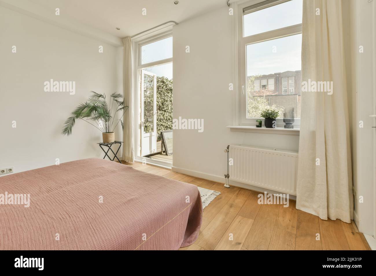 Komfortables Bett und Lampe in einem kleinen, schmalen Schlafzimmer im minimalistischen Stil mit weißen Wänden und schwarzen Vorhängen am Fenster in einem modernen Apartment Stockfoto