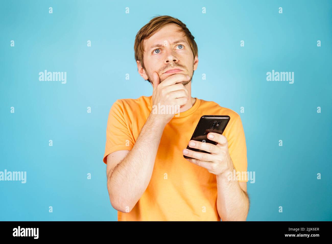 Porträt eines nachdenklichen, nachdenklichen Mannes, der Smartphone in einer Hand hält und das Kinn auf blauem Hintergrund berührt. Stockfoto