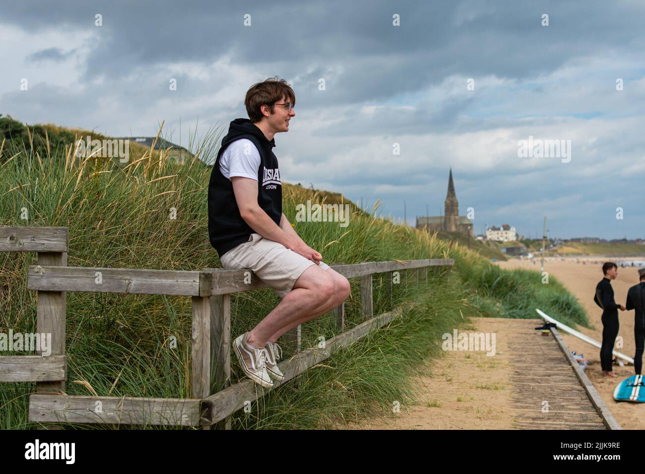 Natürliches Porträt eines jungen Mannes, der auf einem Zaun auf der Promenade am Strand von Longsands in Tynemouth sitzt, mit der Kirche im Hintergrund Stockfoto