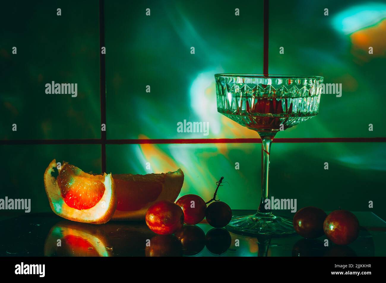 Kristallglas für Cocktails auf einer Glasoberfläche. Früchte - Trauben, Grapefruit neben dem Glas. Abstrakter Hintergrund. Stockfoto