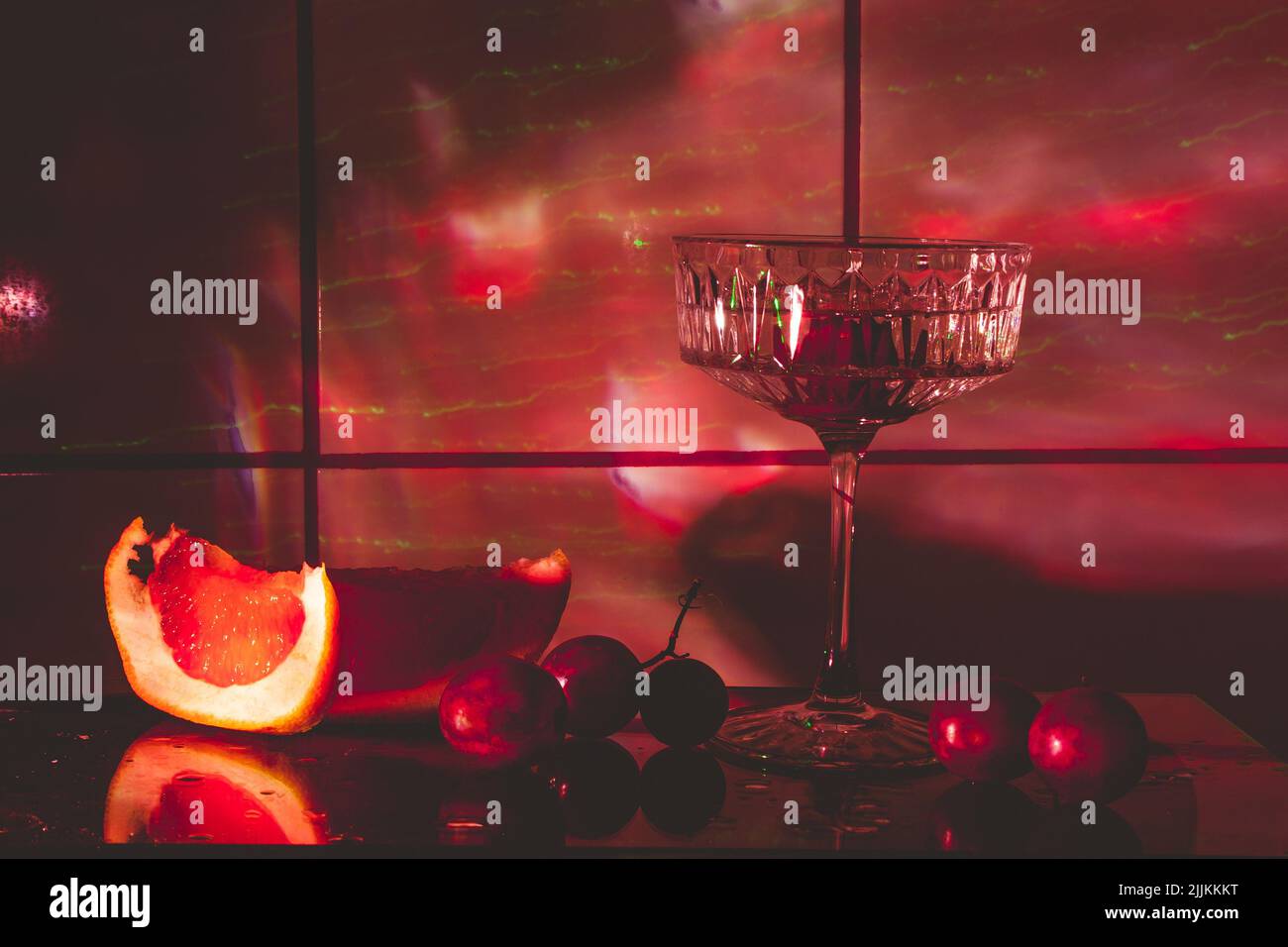Kristallglas für Cocktails auf einer Glasoberfläche. Früchte - Trauben, Grapefruit neben dem Glas. Abstrakter Hintergrund. Stockfoto