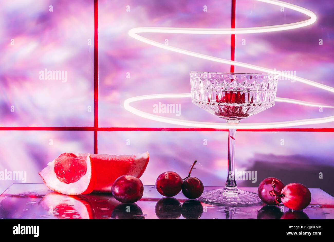 Kristallglas für Cocktails auf einer Glasoberfläche. Früchte - Trauben, Grapefruit neben dem Glas. Abstrakter Hintergrund. Stockfoto