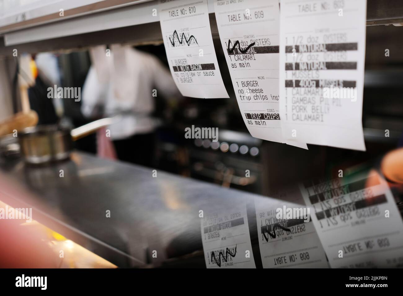 Check-on, Ticketbestellungen auf dem Pass einer professionellen Küche mit Köchen Kochen im Hintergrund. Menüpunkte, die von Kellnern in der Reihenfolge ihrer Priorität organisiert werden. Stockfoto